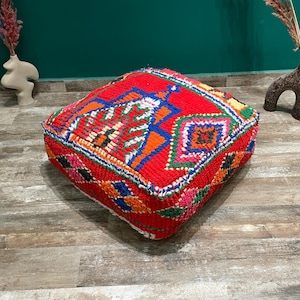Könnte beinhalten: Ein quadratischer, roter marokkanischer Pouf mit einem lebendigen geometrischen Muster in Blau, Orange, Grün und Rosa. Der Pouf besteht aus gewebtem Stoff und steht auf einem Holzboden. Dekorative Vasen sind im Hintergrund.