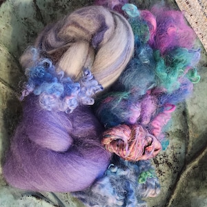 May include: A collection of colorful wool roving in shades of purple, blue, and green. The roving is arranged in a bowl and ready for spinning or felting.