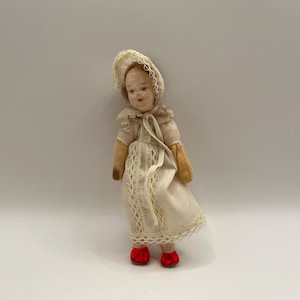 May include: A small vintage doll with a painted face, wearing a cream-colored fabric bonnet and a matching dress with lace trim detailing. The doll has tan-colored arms and is wearing bright red shoes on its feet, standing in an upright position.