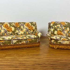 May include: A miniature sofa and matching armchair upholstered in a cream fabric featuring a yellow rose and green vine floral pattern. Both pieces of dollhouse furniture are finished with a decorative gold-toned fringe trim along the base.