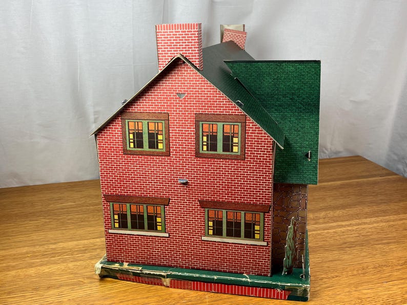 May include: A vintage-style toy dollhouse constructed from printed cardboard, featuring a red brick exterior pattern, a dark green roof, and a prominent chimney. The structure includes four rectangular windows with green frames and yellow interior panes.