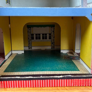 May include: A cardboard dollhouse interior featuring yellow walls, a teal floor, and a printed window with curtains. The structure includes arched doorways, a red brick pattern on the exterior side, and a blue horizontal beam across the top.