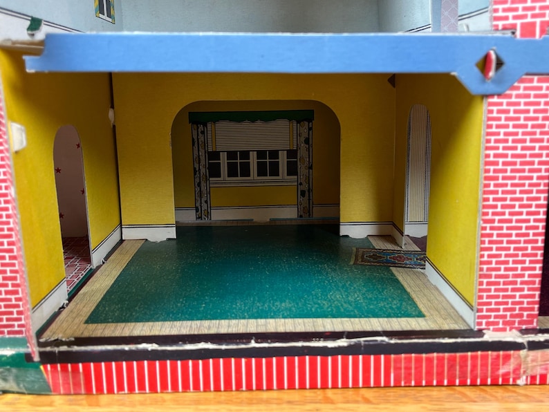 May include: A cardboard dollhouse interior featuring yellow walls, a teal floor, and a printed window with curtains. The structure includes arched doorways, a red brick pattern on the exterior side, and a blue horizontal beam across the top.