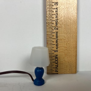 May include: Miniature blue table lamp with a white lampshade. The lamp is approximately 1.5 inches tall. A wooden ruler is next to the lamp, and a brown electrical cord extends from the lamp.