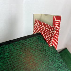 May include: A miniature toy house roof section featuring a dark green shingled pattern and a tall, rectangular chimney with a red and white brick design. The structure is made of flat, printed material with sharp edges and a visible interior corner.