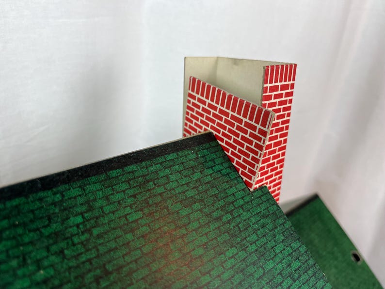 May include: A miniature toy house roof section featuring a dark green shingled pattern and a tall, rectangular chimney with a red and white brick design. The structure is made of flat, printed material with sharp edges and a visible interior corner.