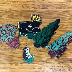 May include: A collection of five die-cut paper ephemera pieces featuring a potted plant with red flowers, a black vintage baby carriage with a yellow hood and swan graphic, a small cat in a green box, and two stylized evergreen tree cutouts.