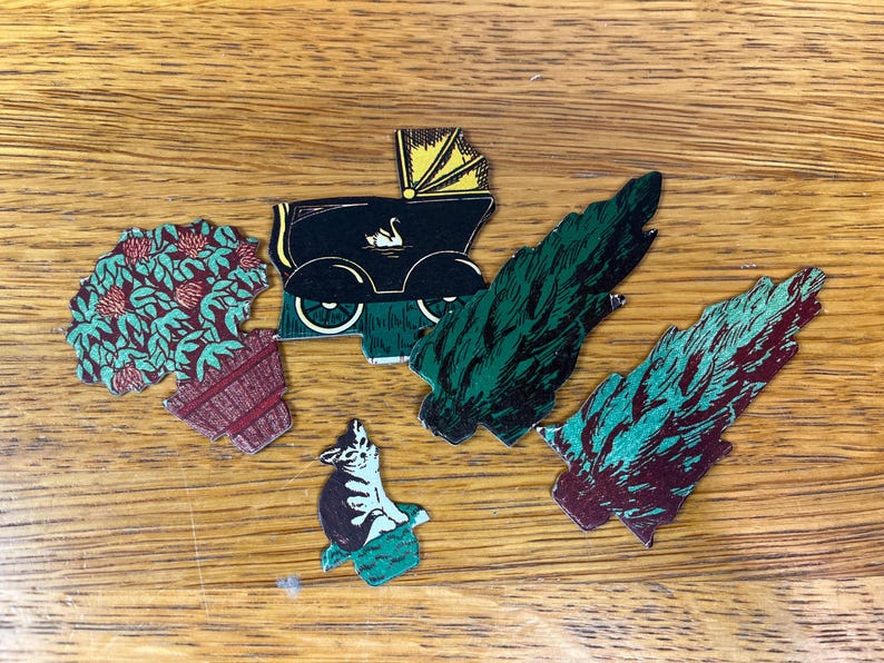 May include: A collection of five die-cut paper ephemera pieces featuring a potted plant with red flowers, a black vintage baby carriage with a yellow hood and swan graphic, a small cat in a green box, and two stylized evergreen tree cutouts.