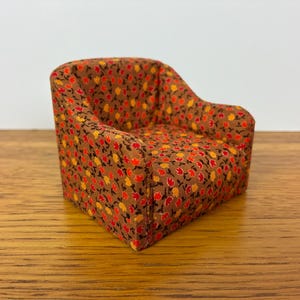 May include: A miniature armchair upholstered in a brown fabric featuring a dense, repeating pattern of small red and yellow tulip-like flowers. The chair has a rounded backrest, integrated armrests, and a compact, low-profile seat design.