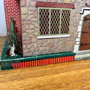 May include: A vintage tin toy house featuring a stone-textured facade with a brown wooden door, a green-framed lattice window, and a red brick side wall. A small illustration of a cat sits near a green tree on the dark green base of the structure.