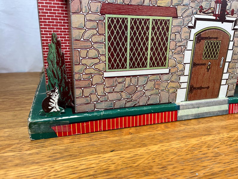 May include: A vintage tin toy house featuring a stone-textured facade with a brown wooden door, a green-framed lattice window, and a red brick side wall. A small illustration of a cat sits near a green tree on the dark green base of the structure.
