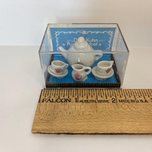 May include: A miniature porcelain tea set in a clear plastic box. The set includes a teapot, creamer, and two teacups and saucers. The tea set is decorated with pink roses. The box has a blue background with the text "D. Kuhn & R. Rebel" printed on it.