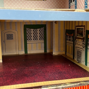May include: A dollhouse interior featuring a room with yellow and blue striped wallpaper, a dark red floor, and a white door. The walls include printed details of a fireplace, green curtains, and a window with a lattice pattern design.