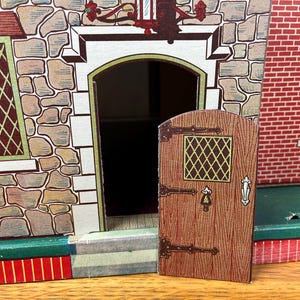 May include: A toy house facade featuring a stone-textured wall, a red brick section, and a hinged wooden-style door with a small diamond-patterned window. The structure includes a green base, a dark doorway, and detailed illustrations of windows.