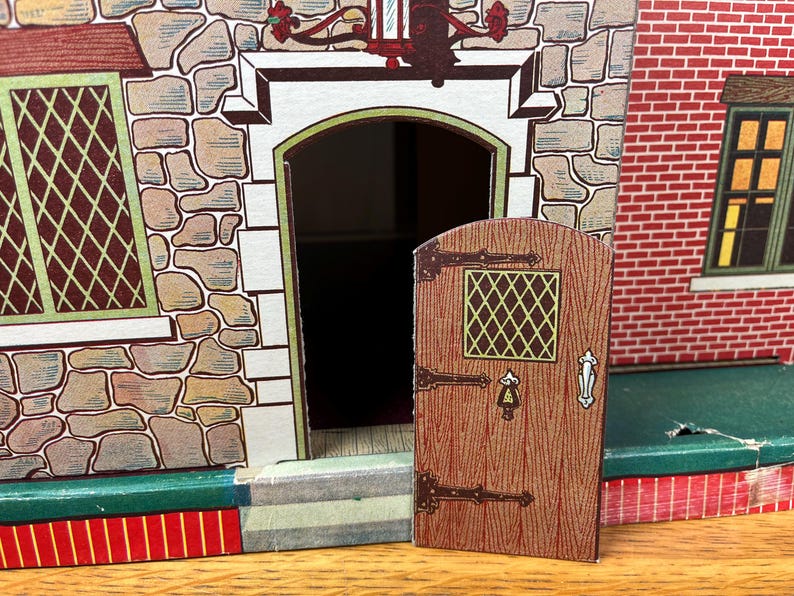 May include: A toy house facade featuring a stone-textured wall, a red brick section, and a hinged wooden-style door with a small diamond-patterned window. The structure includes a green base, a dark doorway, and detailed illustrations of windows.