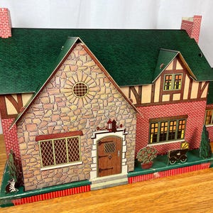 May include: A vintage-style toy dollhouse featuring a green shingled roof, stone-patterned front facade, and red brick side walls. The structure includes a brown front door, multiple windows with lattice detailing, and decorative printed shrubbery.