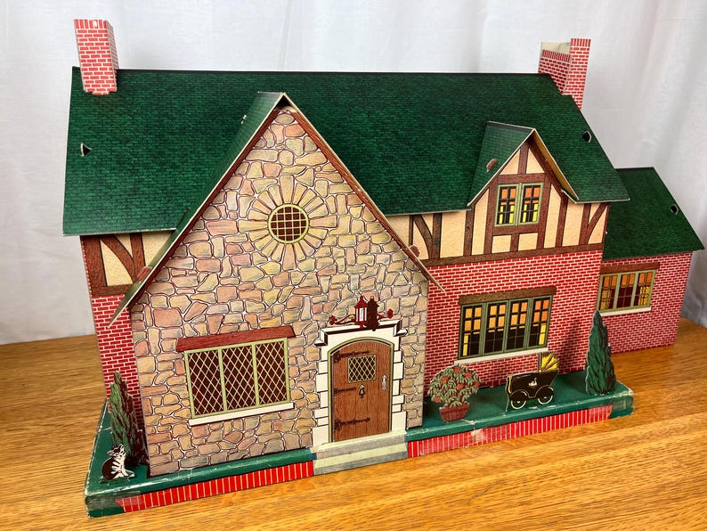 May include: A vintage-style toy dollhouse featuring a green shingled roof, stone-patterned front facade, and red brick side walls. The structure includes a brown front door, multiple windows with lattice detailing, and decorative printed shrubbery.