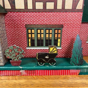 May include: A vintage cardboard toy house facade featuring a red brick wall, a stone-textured side section, and green-framed windows. In front, a small black baby carriage with a yellow hood and a swan graphic sits between a potted bush and a tree.