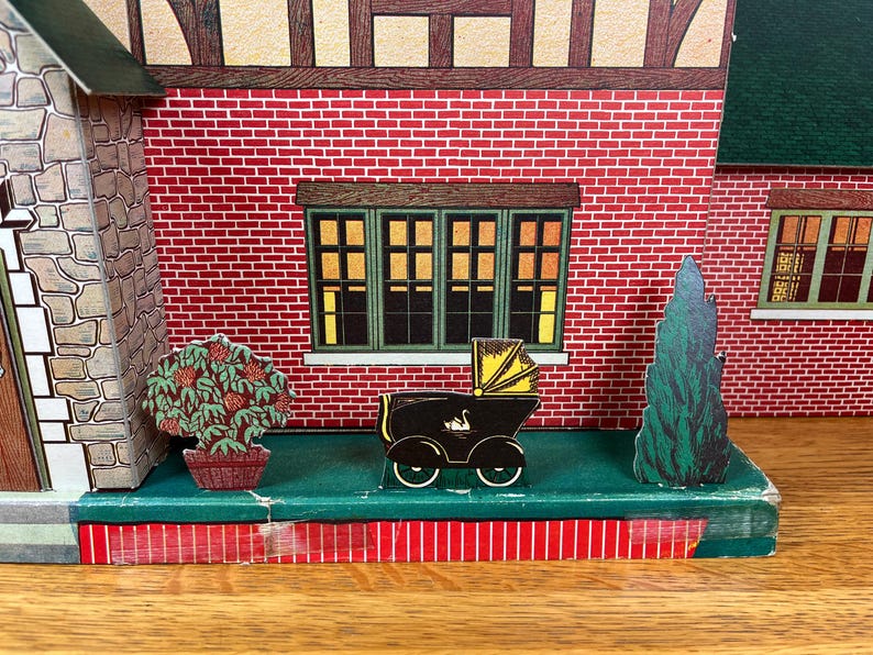 May include: A vintage cardboard toy house facade featuring a red brick wall, a stone-textured side section, and green-framed windows. In front, a small black baby carriage with a yellow hood and a swan graphic sits between a potted bush and a tree.