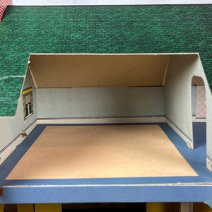 May include: A cardboard dollhouse featuring a green shingled roof and red brick exterior walls. The interior room has light-colored walls, a blue floor border, and a central beige floor area. A window with a yellow frame is visible on the left wall.