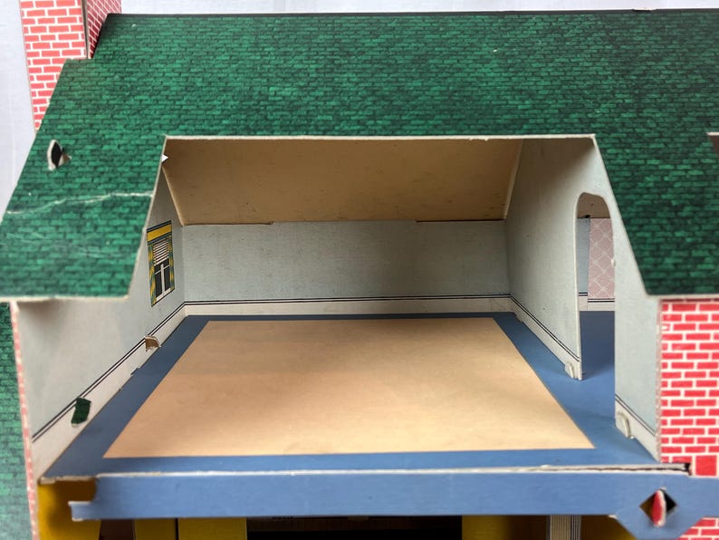 May include: A cardboard dollhouse featuring a green shingled roof and red brick exterior walls. The interior room has light-colored walls, a blue floor border, and a central beige floor area. A window with a yellow frame is visible on the left wall.