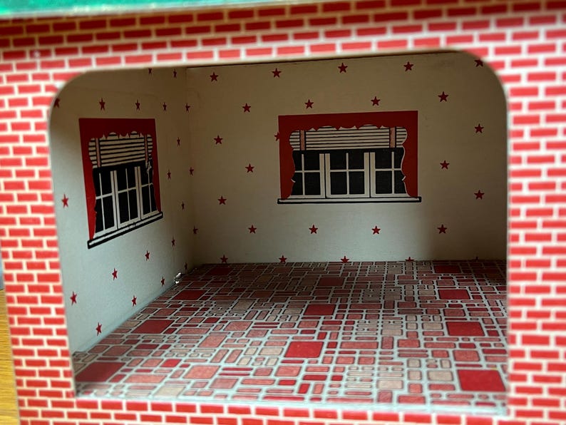 May include: A dollhouse room interior featuring white walls with a red star pattern, two windows with red valances and blinds, and a floor covered in a red and tan geometric tile pattern. The exterior frame is printed with a red brick design.