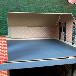 May include: A cardboard dollhouse featuring a green shingled roof and red brick exterior walls. The interior attic room has light pink wallpaper with a white diamond pattern, blue flooring, and two windows with blue and white polka dot curtains.