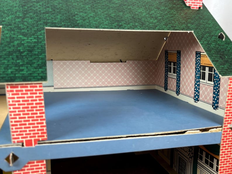 May include: A cardboard dollhouse featuring a green shingled roof and red brick exterior walls. The interior attic room has light pink wallpaper with a white diamond pattern, blue flooring, and two windows with blue and white polka dot curtains.