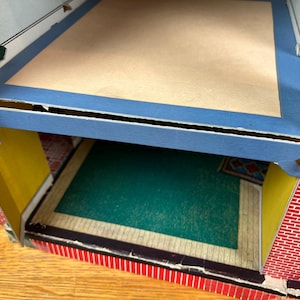 May include: A multi-level cardboard dollhouse structure featuring a red brick pattern on the exterior walls, yellow interior wall panels, a blue floor border, a tan upper floor, and a green floor section on the lower level with a small rug.