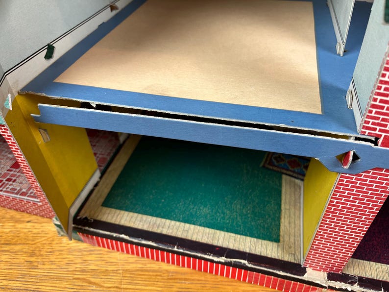 May include: A multi-level cardboard dollhouse structure featuring a red brick pattern on the exterior walls, yellow interior wall panels, a blue floor border, a tan upper floor, and a green floor section on the lower level with a small rug.