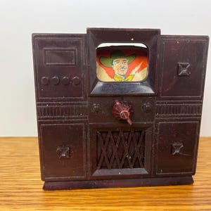 May include: A dark brown plastic toy television set featuring a screen with a colorful illustration of a person wearing a cowboy hat. The front panel includes molded knobs, a central dial, and a speaker grille with a cross-hatched pattern design.