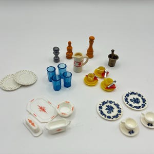 May include: A collection of miniature kitchen and dining items including white plates with lace-like edges, blue translucent tumblers, a white pitcher with red floral patterns, yellow cups, a mortar and pestle, and various white ceramic dishes.