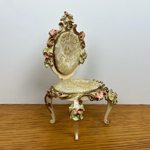 May include: A miniature decorative chair with a cream-colored frame and floral accents. The chair features a patterned seat and backrest, with gold trim and pink and green floral embellishments. The legs are curved and delicate.