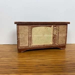 May include: A miniature vintage television set made of wood. The TV has a brown frame with a light-colored screen and speaker panels on either side. It features knobs and a retro design, evoking a mid-century aesthetic.