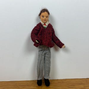 May include: A small doll with brown hair, wearing a dark red textured sweater over a white collared shirt with a small green, white, and red striped tie. The doll has light gray trousers and black shoes, featuring a painted face with brown eyes.
