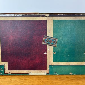 May include: A rectangular cardboard box with a worn exterior featuring a large dark maroon panel on the left and a green panel on the right, both framed by tan borders. A small rectangular label with a blue, red, and gold floral pattern is centered.