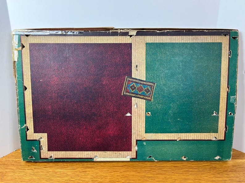 May include: A rectangular cardboard box with a worn exterior featuring a large dark maroon panel on the left and a green panel on the right, both framed by tan borders. A small rectangular label with a blue, red, and gold floral pattern is centered.