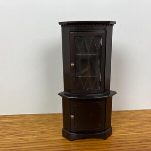 May include: A dark brown miniature corner cabinet featuring a top section with a glass door displaying a diamond-shaped grid pattern and a solid bottom cabinet door. Both doors are equipped with small, round, metallic-colored knobs.