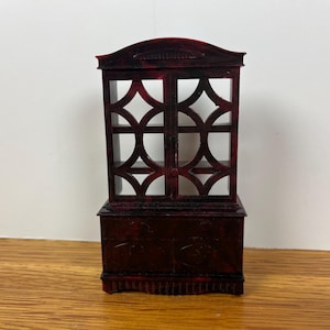 May include: A miniature, dark red cabinet with a decorative, openwork design on the upper doors. The cabinet has a rectangular base and a curved top. The wood grain is visible. The cabinet is a decorative item.