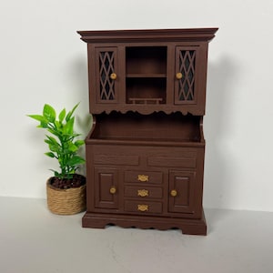 May include: A miniature brown wooden hutch with two upper cabinets featuring diamond-patterned glass inserts and three lower drawers with gold-colored hardware. A small potted plant sits to the left.
