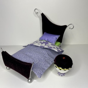 May include: A miniature purple velvet bed with a silver frame and a floral patterned comforter. The bed has a purple pillow and a small black velvet footstool with a silver trim.