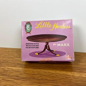 May include: A vintage box for "Little Hostess" miniature furniture by Marx. The box is lavender with an illustration of a brown table with gold accents. Text includes "Distinctive Miniature Furniture".