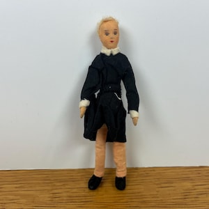 May include: A fabric doll with blonde hair and a painted face, wearing a black long-sleeved coat with a white collar and white cuffs. The doll has light peach-colored fabric legs and wears small black shoes. The doll stands in an upright position.