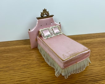 May include: A miniature pink bed with a decorative headboard and two pillows. The bed frame is trimmed with gold and has a white lace skirt. The headboard is topped with a decorative element.