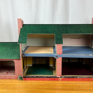 May include: A two-story cardboard dollhouse featuring a green shingled roof and red brick exterior walls. The structure includes an attached garage, open-front rooms with printed interior wallpaper, and blue and yellow floor sections throughout.