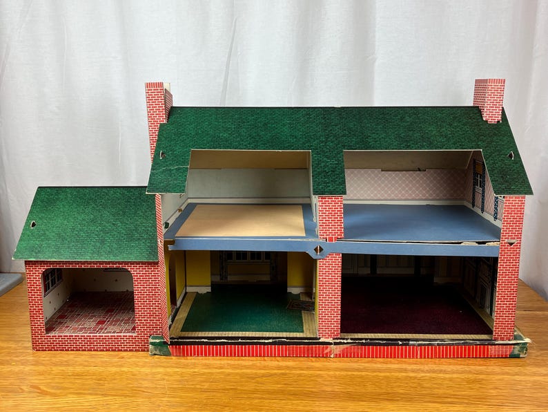 May include: A two-story cardboard dollhouse featuring a green shingled roof and red brick exterior walls. The structure includes an attached garage, open-front rooms with printed interior wallpaper, and blue and yellow floor sections throughout.