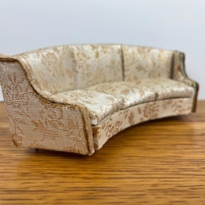 May include: A miniature curved sofa with a cream-colored floral pattern. The sofa has a curved back and armrests, and the fabric has a gold floral design. The sofa is a dollhouse or miniature furniture item.