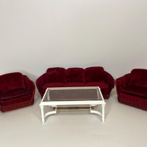 Puede incluir: Un conjunto de sala de estar con un sofá de terciopelo rojo y dos sillones a juego. Una mesa de centro blanca con tapa de cristal se encuentra en el centro del conjunto.