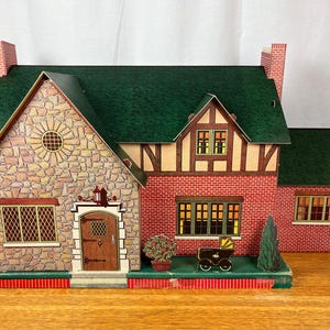 May include: A toy dollhouse featuring a green roof, a stone-patterned facade on the left section, and red brick-patterned walls on the right. The structure includes a brown front door, multiple windows with lattice details, and a small toy carriage.