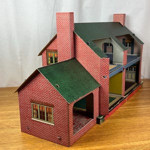 May include: A vintage-style dollhouse featuring a red brick exterior pattern, a dark green gabled roof, and two prominent chimneys. The structure is open on one side, revealing multiple interior rooms with colored walls and printed window details.